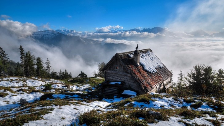 Pequeña casa con un poco de nieve encima de ella rodeada de niebla cerca de unas montañas 