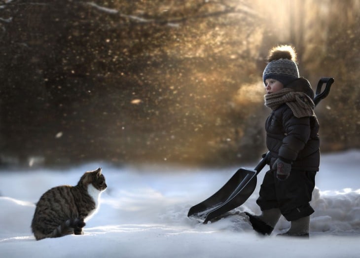 niño con una pala juntando nieve al lado del gato