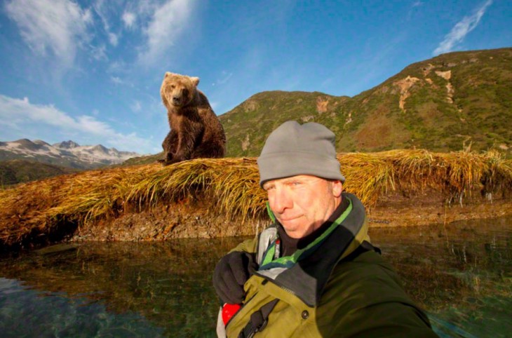 Hombre tomándose una Selfie en un lago con un osos detrás de él 