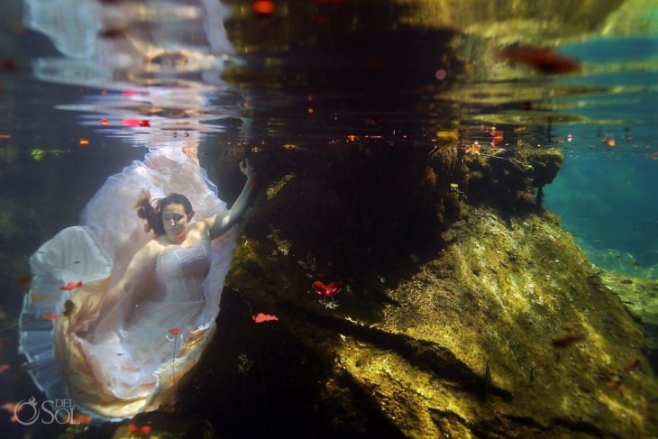 una mujer vestida de novia posando debajo del mar sosteniéndose de una piedra 