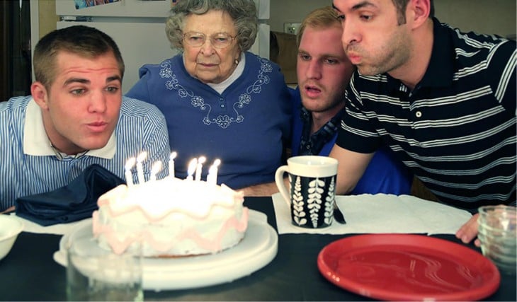 joven soplando las velas de su pastel de cumpleaños acompañado de sus otros dos hermanos y abuela