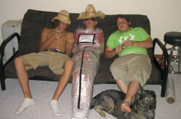 3 chicos en un sofá, el del medio, envuelto en cinta