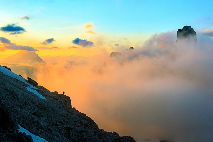 Persona a lo lejos a la orilla de una piedra de una montaña observando hacia otras montañas 