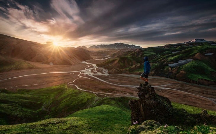 Hombre con vestimenta deportiva parado en una pequeña montaña viendo como sale el sol de otras montañas a su alrededor 