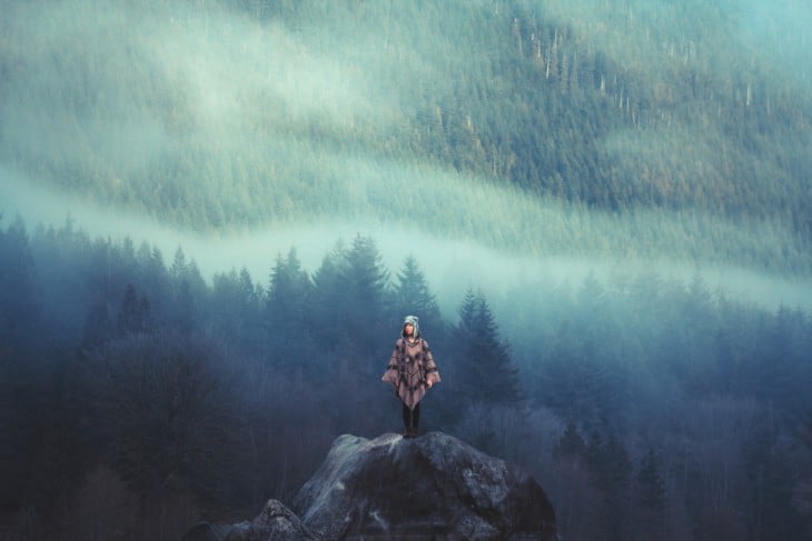 Chica parada sobre una piedra y al fondo un gran bosque repleto de pinos 