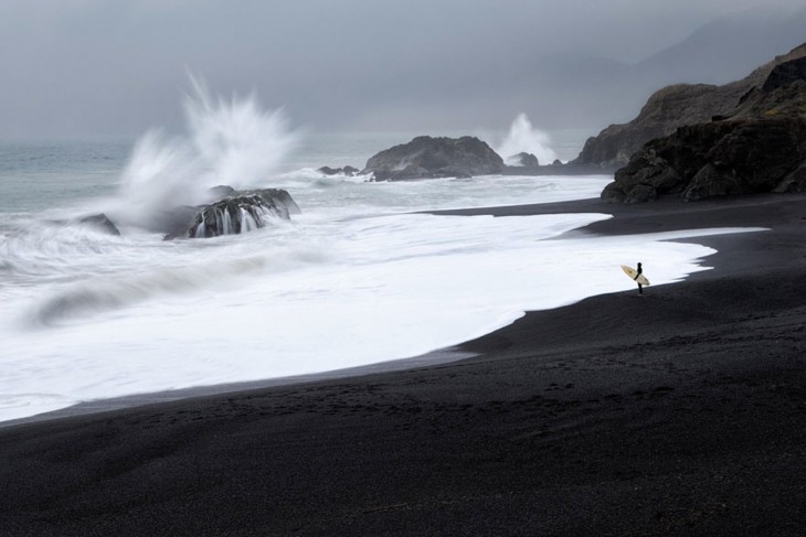 Hombre con una tabla de Surf parado a la orilla de un mar donde las olas chocan con las rocas 