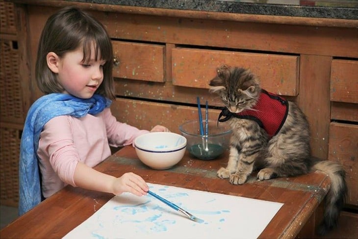 Niña frente a una mesa con un pincel en la mano pintando sobre una hoja y un gato a su lado 