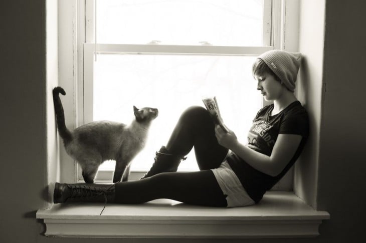 Chica acostada en una ventana y un gato frente a ella 