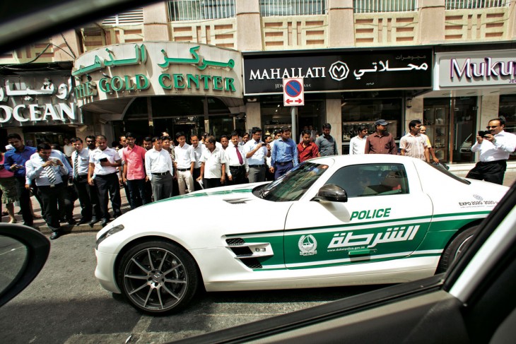 Patrulla pasando por la calle y personas a la orilla de una calle observando los carros deportivos 