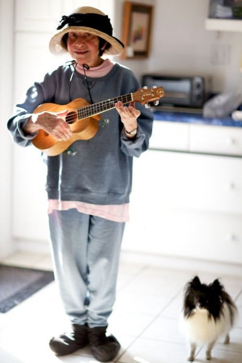 Mujer anciana tocando la guitarra y un perro a un costado de ella 