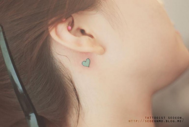 Tatuaje minimalista de un corazón verde detrás del oído de una chica 
