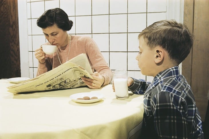 Madre con un periódico en sus manos a lado de su hijo que toma leche con galletas 
