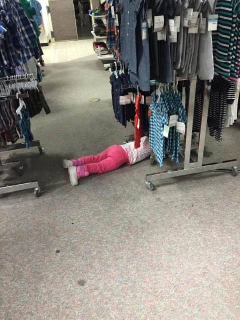 niña en el suelo entre ropa colgada en un centro comercial 