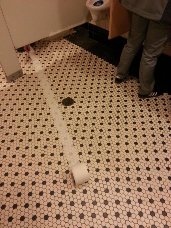 Persona dentro de un baño público intentando regresar el papel que se rodo por el piso 