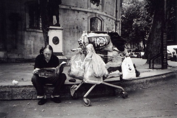 Fotografía de un vagabundo sentado en el suelo con un carro lleno de bolsas en un lado 