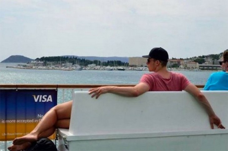 Chico sentado en una banca frente a un puerto que parece tener piernas de mujer 