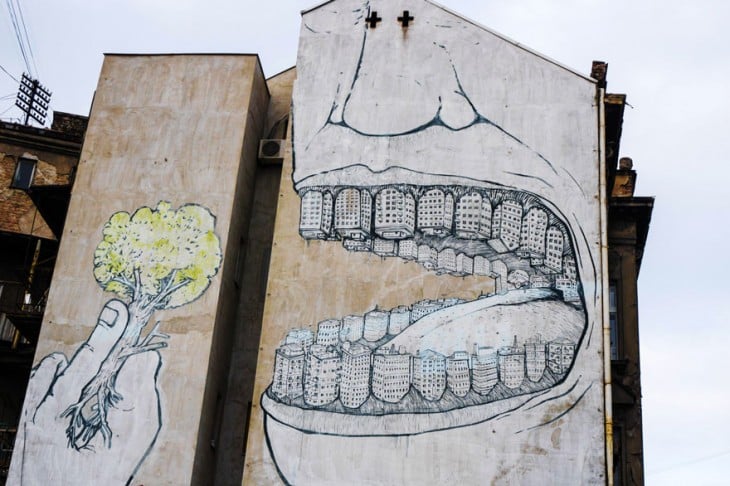 Grafiti de una boca con dientes de edificios consumiéndose un árbol 