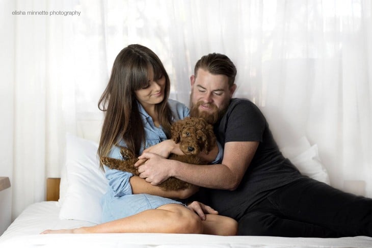Un matrimonio recostados en la cama sosteniendo en sus brazos a su pequeño y adorable cachorro 