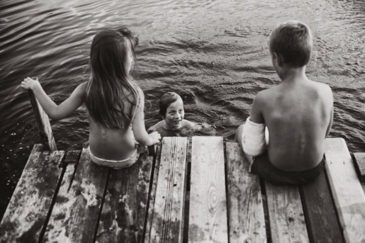 una niña y un niño sentados en un puente viendo a un niño que esta dentro de un lago 