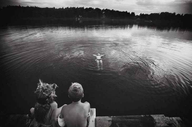 niño nadando en un lago mientras dos niños sentados en un muelle lo ven 