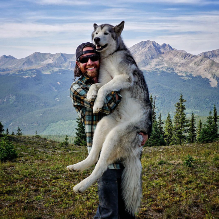 Kelly Lund cargando a su perrolobo Loki en un paisaje natural cerca de unas montañas 