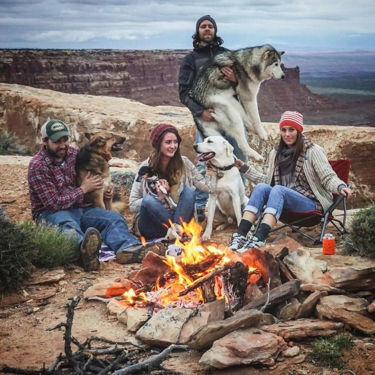 Grupo de personas con sus perros frente a una fogata en un paisaje con montañas y un poco frío 