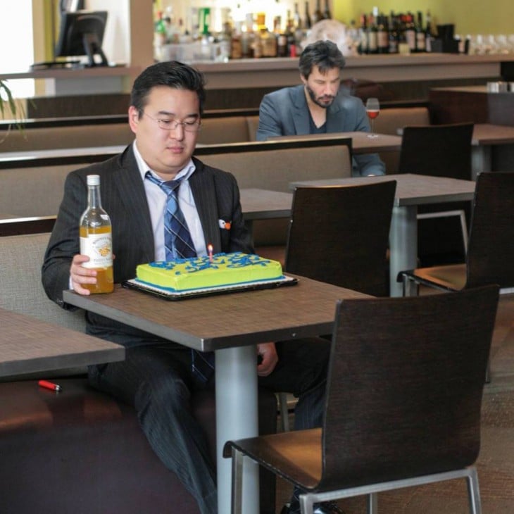 Fotografía de un chico sentado frente a la mesa de un bar con un pastel de cumpleaños y una botella en su mano con una persona detrás de él 