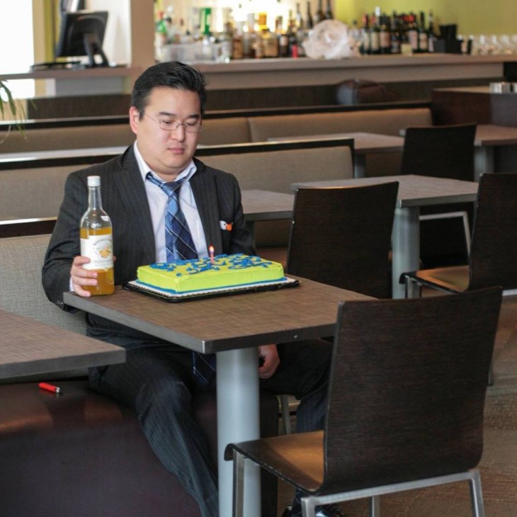Fotografía de un chico sentado frente a la mesa de un bar con un pastel de cumpleaños y una botella en su mano 