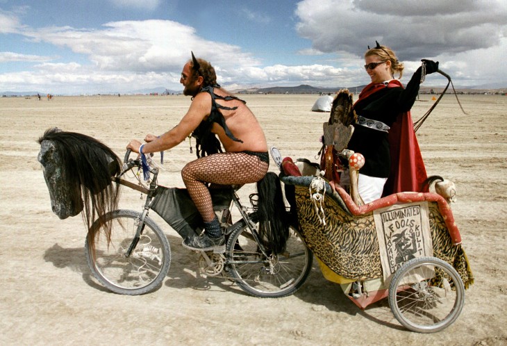 Un hombre en bicicleta estirando a una mujer sobre un carro con un látigo en el festival Burning Man 