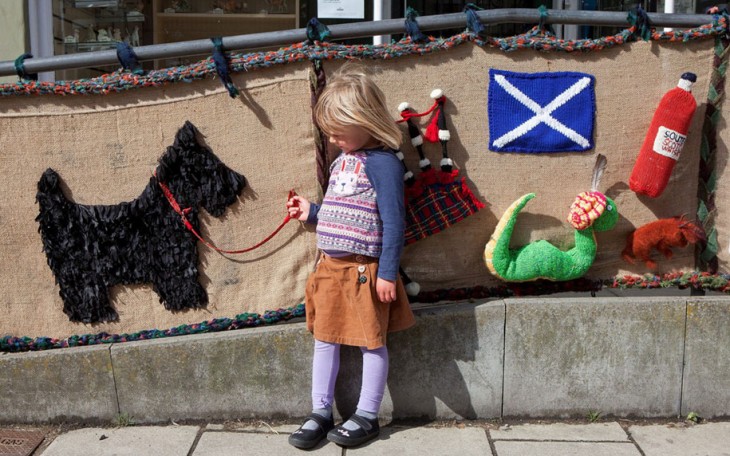 Niña sosteniendo en su mano la correa de un perro que esta tejido en una valla de una calle en Escocia 