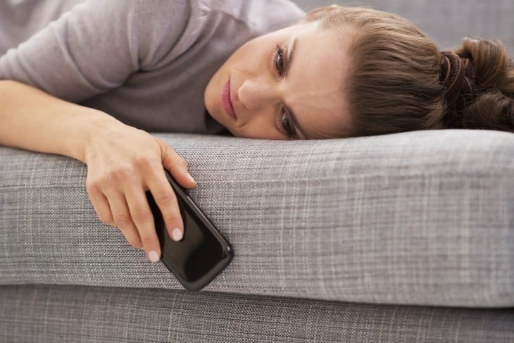 una chica recostada en un sillón con cara de depresión y con el celular en la mano hacia abajo