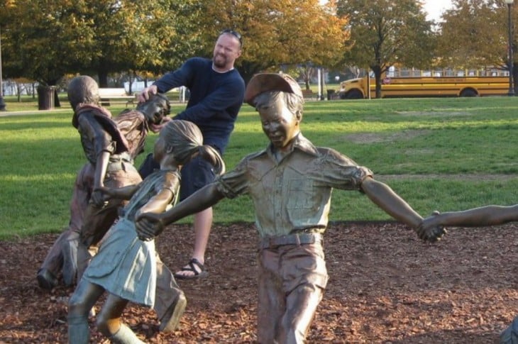 fotografía de una estatua de unos niños jugando a la ronda mientras un hombre simula estar agarrando la cabeza de una estatua