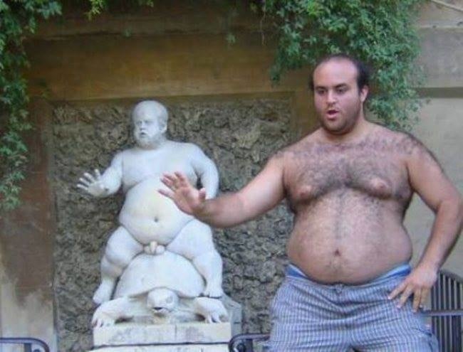 hombre sin camisa imitando la pose de una pequeña estatua en la pared 