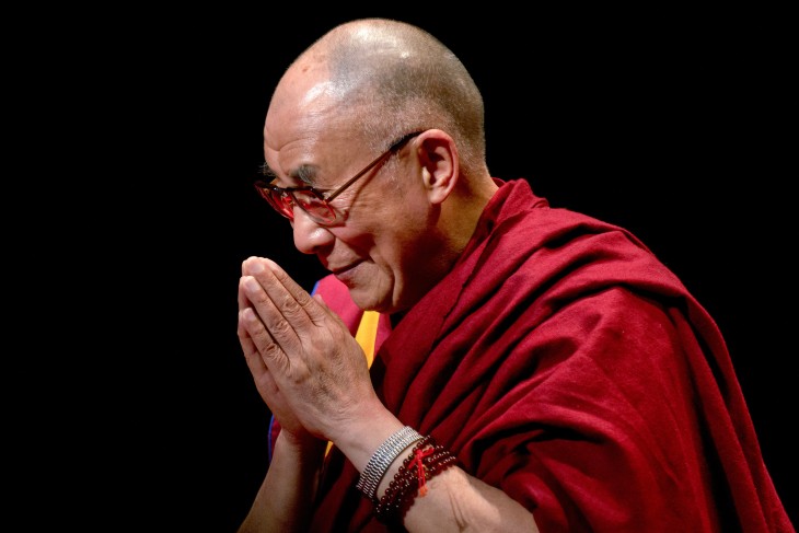 Dalai Lama con la cabeza un poco inclinada con sus manos juntas haciendo oración 