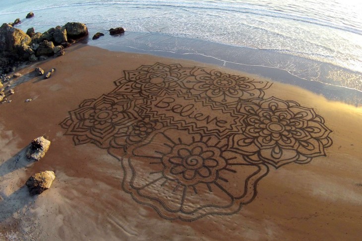 Dibujo de flores con la palabra "Believe" en la arena sobre una playa 