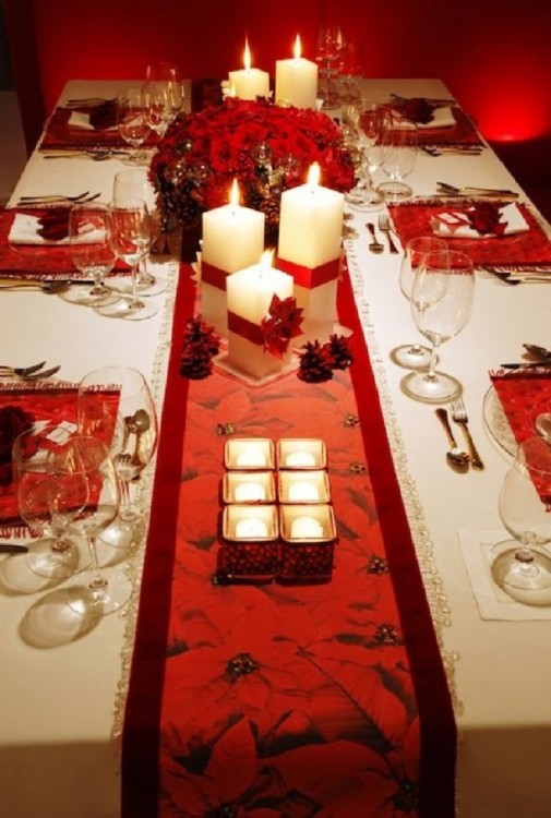 Mesa adornada con velas pequeñas y veladoras grandes cuadradas y en el centro un arreglo de flores en color rojo 