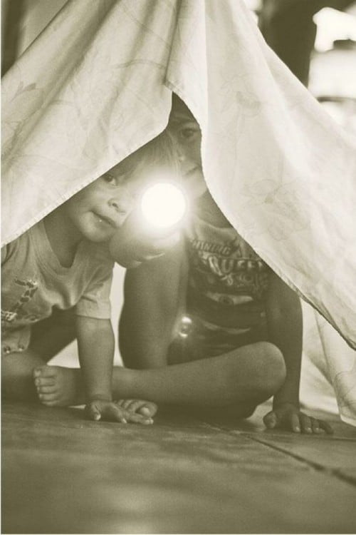 Dos niños debajo de una sabana con una linterna en la mano 