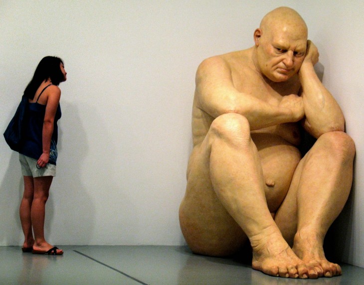 Escultura gigante de un hombre sentado en la esquina de un cuarto 