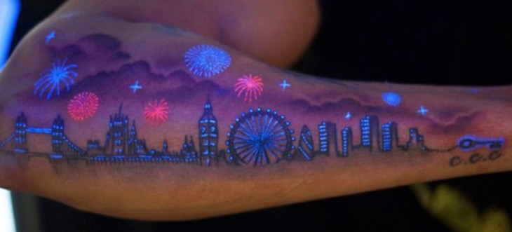 Tatuaje ultravioleta en un brazo con el diseño de una ciudad con edificios y pirotecnia 