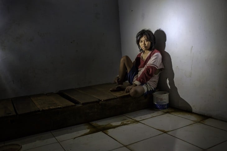 Fotografía de una chica sentada en una base de madera recargada en la pared en un hospital mental en Indonesia