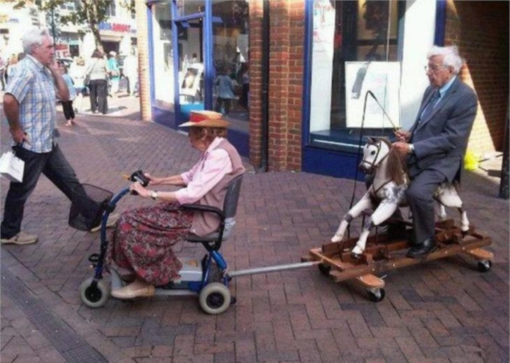 Abuela con su silla de ruedas mecánica y con ella jala un carrito en donde va un caballito de juguete y en él va montado el abuelo