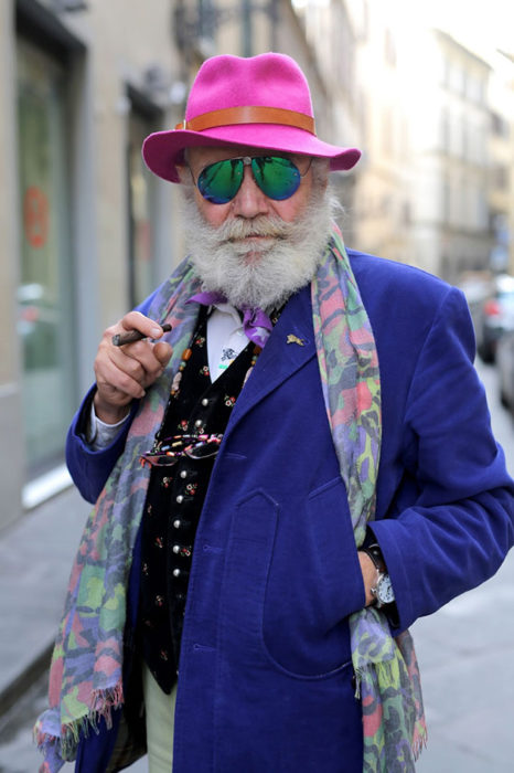 Hombre de +60 años con saco azul rey, sombero rosa, mascada, chaleco negro de colores, lentes y puro