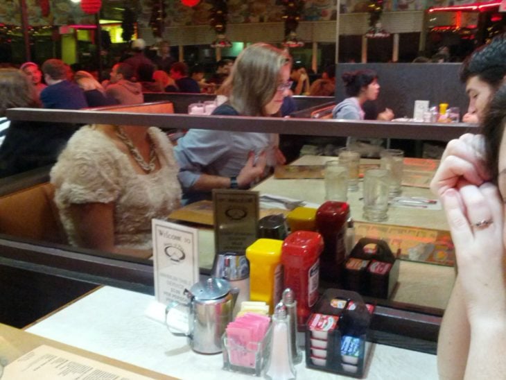 Fotografía engañosa en donde, por un espejo que divide dos mesas de un restaurante, parece que la mujer que come no tiene cabeza