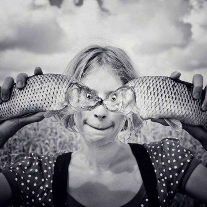 Mujer sosteniendo dos pescados de perfil haciendo la ilusión de que los ojos de los pescados son de ella