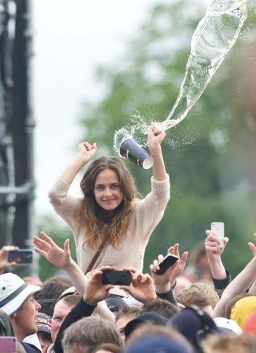 Una mujer en un concierto sonríe para la cámara antes de que un vaso con una bebida la golpee en la cabeza