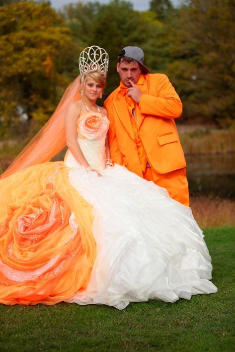 Novia con el velo naranja, corona de reina y en su vestido tiene cosas naranjas simulando una flor. El novio va vestido todo de naranja con una gorra gris de lado