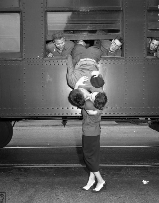 Amor en tiempos de guerra. dos soldados sostienen las piernas de otro que sale de la ventana de un camión para besar a la mujer que está abajo