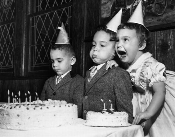 Tres niños frente a dos pasteles de cumpleaños, uno con cara de tristeza, otro soplando las velitas y una más llorando
