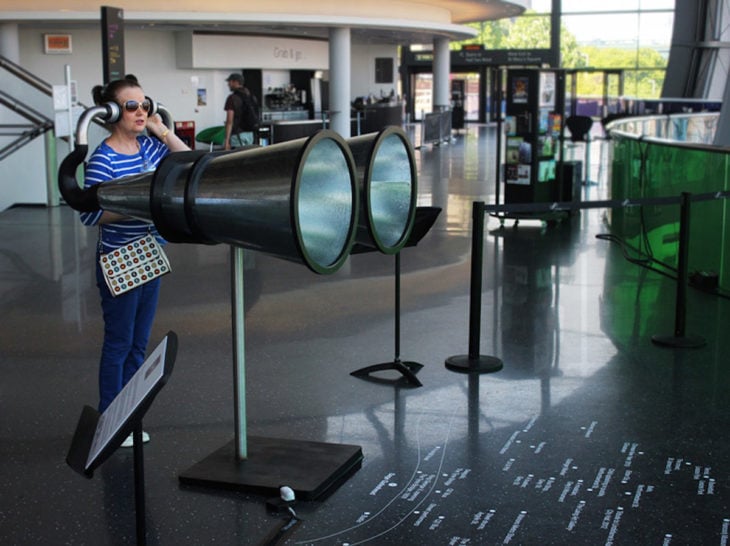 Mujer en lo que parece un aeropuerto escuchando algo en unos binoculares gigantes