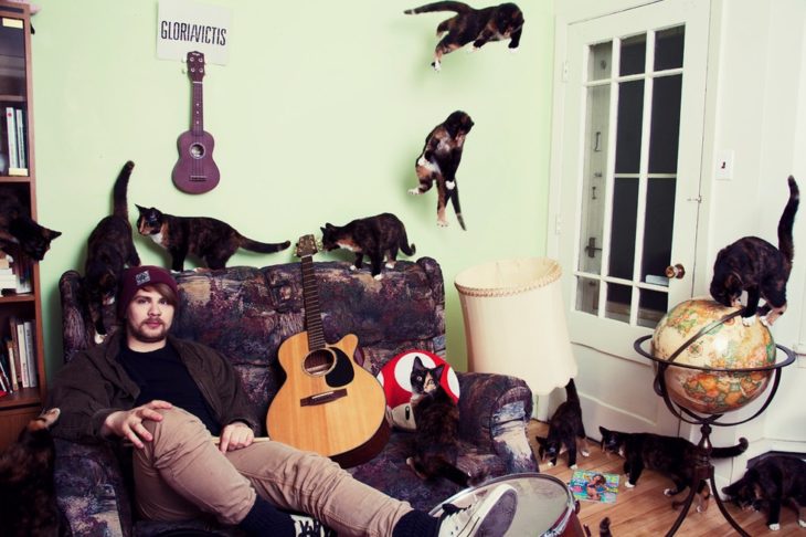 Un chavo en la sala de su casa con su guitarra y sus gatos 